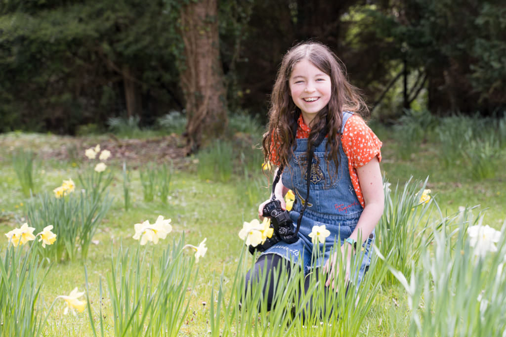 Young girl sitting in the daffodils with her camera and smiling whilst on a photograph course in Sussex