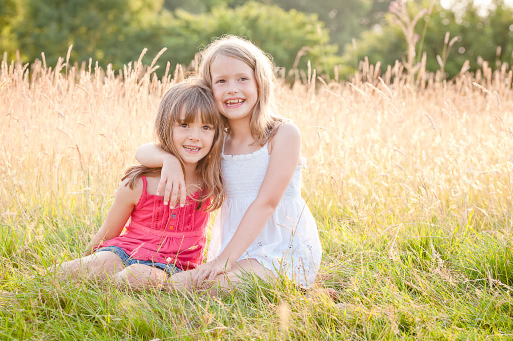 Two sisters sitting in long grass during their summer family photoshoot in Ardingly